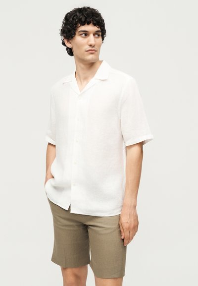 Άνδρας με σγουρά σκούρα μαλλιά που φοράει λευκό κοντομάνικο πουκάμισο με κουμπιά και μπεζ σορτς, στέκεται με το ένα χέρι στην τσέπη.