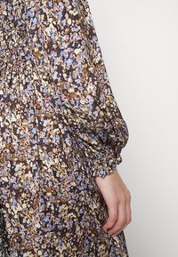 Détail de la manche d'une robe fluide à fleurs marron, bleue et beige avec des poignets élastiques froncés et une main partiellement visible reposant sur le côté.