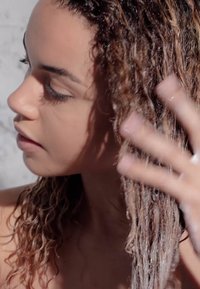 Primer plano de una mujer con cabello rizado y de longitud hasta los hombros en un estado húmedo y espumoso, aplicando un producto para el cabello. Su tono de piel es claro y el fondo es neutro.