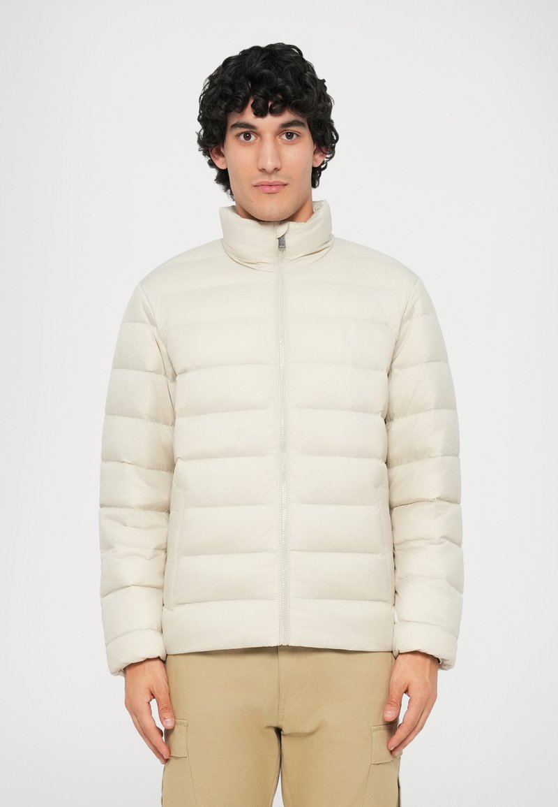 Giacca puffer beige chiaro con baffi orizzontali, colletto alto e chiusura con zip. Texture liscia; presenta un piccolo dettaglio del logo sul petto.