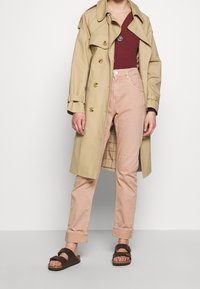 Beige trenčkot se zlatými knoflíky, spárovaný s vínovým ribkovaným topem a růžovými kalhotami s ohrnutými nohavicemi. Hnědé sandály s přezkami.
