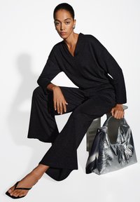 Jumpsuit nero scintillante con gambe larghe e scollatura a V profonda. La modella tiene una borsetta argentata lucida con una texture morbida. Sandali neri piatti visibili.