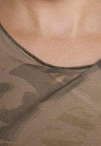 Oliwkowa bluzka w kamuflaż z okrągłym dekoltem i cienkimi przeszyciami wzdłuż brzegów, charakteryzująca się miękką teksturą i warstwowym designem materiału.