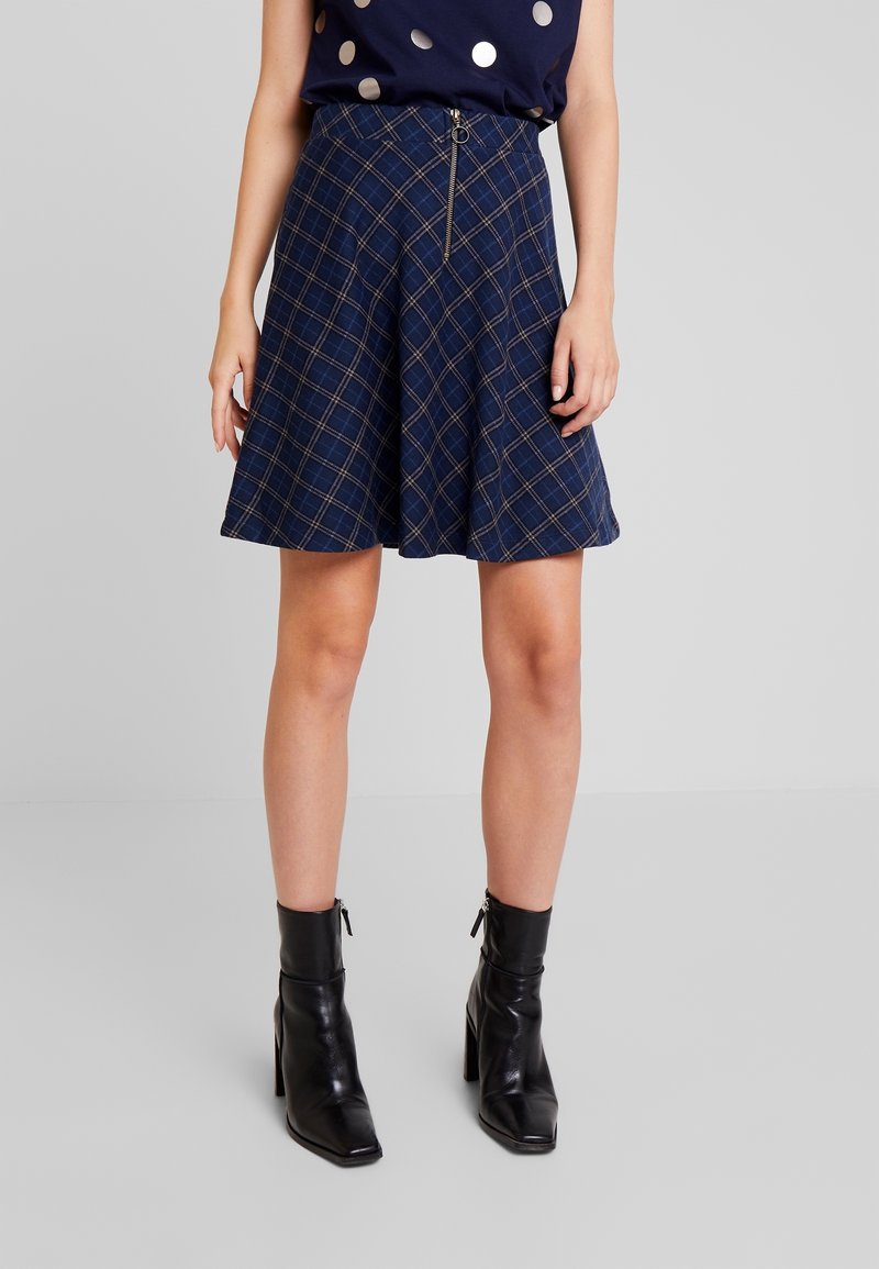 Femme portant une jupe trapèze à carreaux bleus avec une fermeture éclair devant et des bottines noires à talons, debout devant un fond uni.