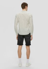 Gestreiftes Langarmshirt in Creme und Marineblau, kombiniert mit dunklen Denim-Shorts. Besitzt hochgerollte Ärmel und aufgesetzte Taschen an der Rückseite, vor einem neutralen Hintergrund.