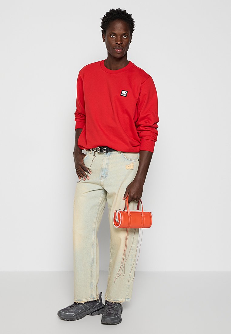 Camicia rossa a maniche lunghe, jeans chiari strappati, sneakers grigie e una piccola borsa arancione dal design strutturato.