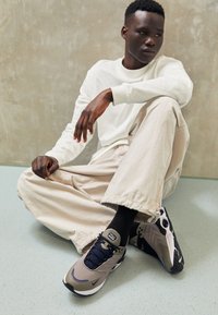 Idrottsskor med en grå och olivfärgad ovandel, marinblå snören och texturerad svart sula. Bärs med ljusbeige byxor och svarta strumpor.