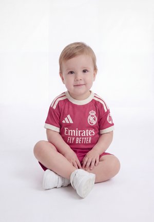 Småbarn sidder med benene i kors iført en rød Real Madrid fodboldtrøje og hvide sneakers mod en ensfarvet hvid baggrund.