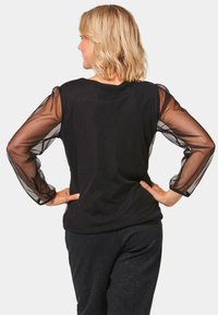 Schwarzes Oberteil mit transparenten, gepufften Ärmeln, rundem Ausschnitt und Rückenfalte. Getragen mit dunklen Glitzerhosen.