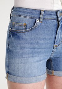 Shorts in denim di colore azzurro chiaro con un classico design a cinque tasche, orli arrotolati e dettagli di cucitura gialla. Texture liscia.