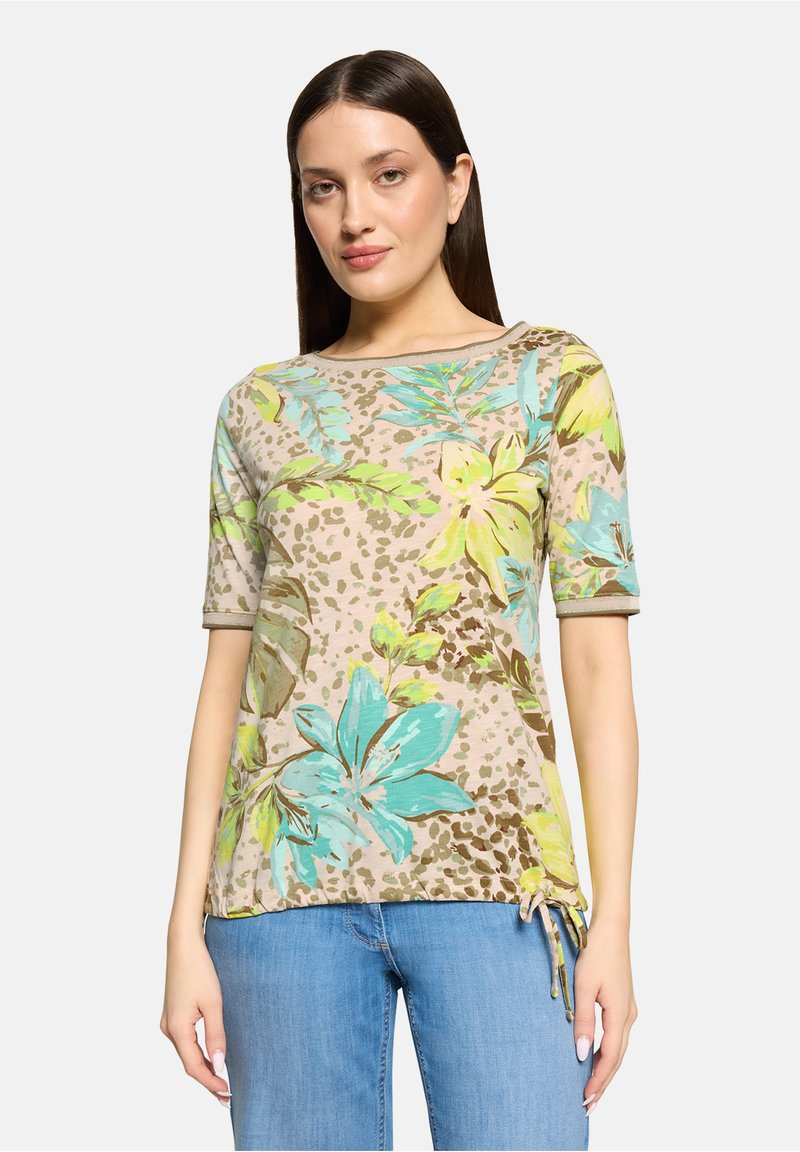 Femme aux longs cheveux foncés portant un haut beige à fleurs bleues et jaunes et un jean bleu clair, debout devant un fond blanc.