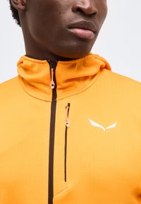 Κοντινό πλάνο ενός ατόμου που φοράει ένα φωτεινό πορτοκαλί μπουφάν με φερμουάρ και ψηλό γιακά, με λευκό μινιμαλιστικό λογότυπο στο στήθος.