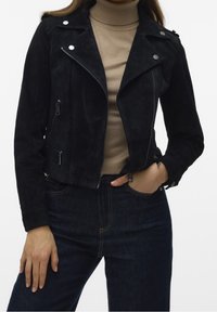 Svart mockajacka med en figursydd design, dragkedjefram, silverfärgad hårdvara och två sidofickor. Bärs över en beige polotröja.