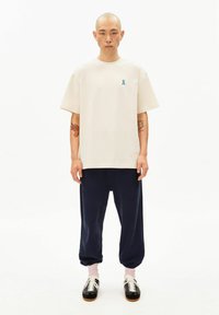 Beige katoenen T-shirt met korte mouwen en een blauw lintlogo, gecombineerd met marineblauwe sweatpants en zwarte sneakers met witte veters.