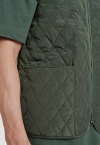 Prošívaná olivově zelená vesta s zipovým uzávěrem a jednou náprsní kapsou, vyrobená z jemné, hladké textilie se diamantovým vzorem.