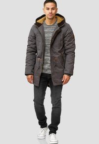 Grijze parka jas met een bruine nep bont voering en accenten, rits- en knoopsluiting, twee voorke pockets, gecombineerd met een gestreepte trui en donkere jeans.