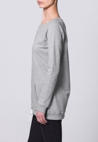 Sudadera gris clara con mangas largas, cuello redondo, bolsillos laterales y dobladillo ajustable con cordón. Tela suave con textura lisa, fit relajado.