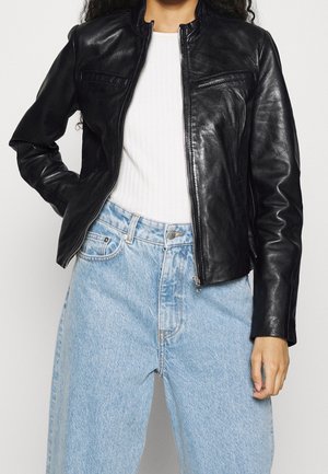 Sort læderjakke med lynlås, høj krave og to vandrette brystlommer. Båret over en hvid ribbet top og lyseblå jeans.
