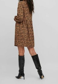 Kobieta w brązowej i czarnej sukience w Tygrysie wzór z długimi rękawami oraz czarnych botkach sięgających do kolan na wysokim obcasie, stojąca na jednolitym tle.