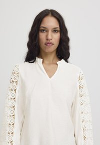 Blusa bianca con scollatura a V e raffinati dettagli in pizzo sulle maniche lunghe. Il tessuto sembra leggero con una finitura texturizzata.