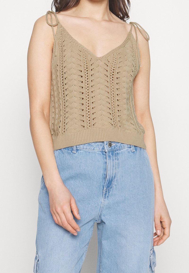 Haut en tricot beige avec décolleté en V, motif ajouré et bretelles à nouer réglables. Associé à un jean bleu clair ample.