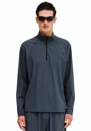 Man met een donkergrijs, lang mouwen, ritsvest en bijpassende broek, met kort donker haar en zwarte wrap-around zonnebril.