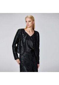 Femme blonde portant une veste texturée noire à fleurs et une jupe assortie sur un top noir en satin à col en V, regardant vers sa gauche sur un fond gris.