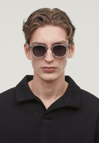 Sonnenbrille mit einem klaren Rahmen und dunklen Gläsern, die eine klassische runde Form aufweisen. Das Modell trägt ein schwarzes Hemd mit Kragen.