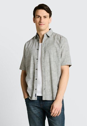 Mężczyzna ubrany w jasnoszarą, krótko rękawową koszulę zapinaną na guziki, na białej koszulce i niebieskich dżinsach, stojący z jedną ręką w kieszeni na jednolitym tle.