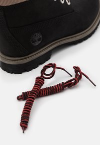 Botte en cuir noir avec col en tissu marron, présentant un logo sur le talon. Lacets en paracorde rouge et noir exposés à côté de la botte.