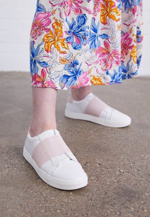 Osoba ubrana w spódnicę w kwiaty w kolorach pomarańczowym, niebieskim i różowym oraz białe wsuwane buty z szerokimi jasnoróżowymi paskami, stojąca na betonowej podłodze.