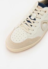 Pantof cu un design sportiv, de culoare albă și bej, cu vârf perforat, accente din suede texturat, șireturi plate și limbă din plasă albastră. Dispune de un emblema pe lateral.