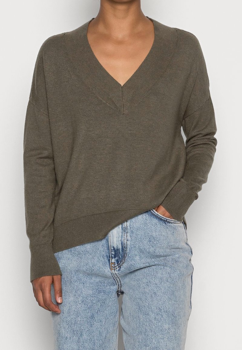 Personne portant un pull à col en V vert olive à manches longues et un jean taille haute bleu clair, une main dans la poche.
