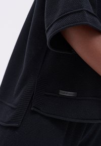 Close-up van een persoon die een gestructureerd zwart breiwerk met korte mouwen draagt, met een klein "blue shadow" label nabij de zoom.
