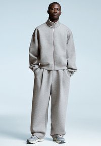 Uomo che indossa una felpa leggera grigia con zip e pantaloni larghi abbinati, insieme a sneakers grigie e bianche, su uno sfondo blu pallido.