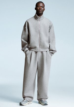 Man wearing light gray zip-up sweatshirt and matching wide-leg pants with gray and white sneakers against pale blue background.