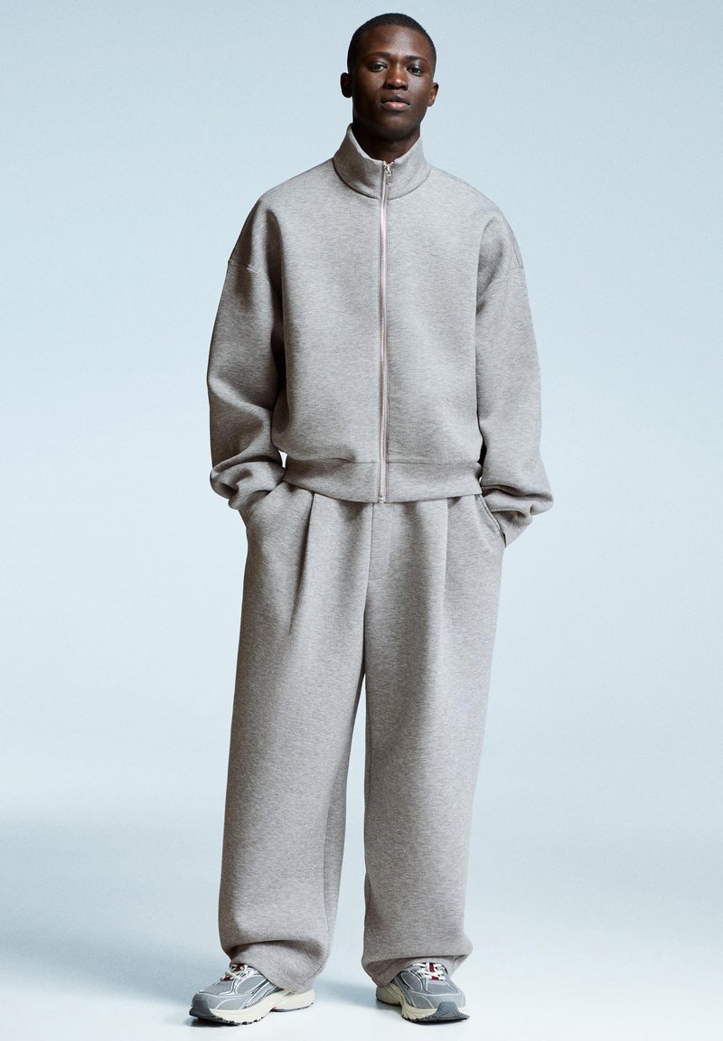 Uomo che indossa una felpa leggera grigia con zip e pantaloni larghi abbinati, insieme a sneakers grigie e bianche, su uno sfondo blu pallido.