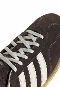 Καφέ σουέντ αθλητικό παπούτσι με λευκό σχέδιο τριών ρίγων, γκρι κορδόνια και υφή σόλας. Διαθέτει στρογγυλεμένη μύτη και ραφές διακοσμητικές.