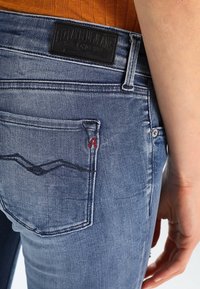 Jeans de mezclilla azul con un acabado desgastado, que presentan un diseño de bolsillo trasero con costuras rojas y una etiqueta de cuero de la marca.