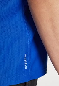 Mėlynas sportinis marškinėlis, pagamintas iš tekstūruoto, drėgmę sugeriančio audinio. Šone turi logotipą su užrašu "AEROREADY".