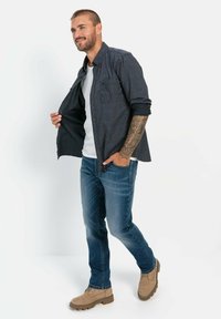 Navy-gepatroneerde overhemd met knopen over een witte t-shirt, blauwe jeans en tan laarzen. Heeft opgerolde mouwen en een ontspannen pasvorm.