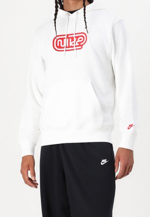 Osoba nosząca białą bluzę z kapturem z czerwonym logo oraz czarne spodnie, oba z logo marki Nike.