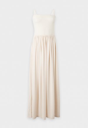 Femme aux longs cheveux foncés portant une robe maxi crème sans manches et des sandales beiges, debout devant un fond clair uni.