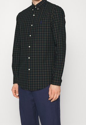 Mężczyzna ubrany w ciemnozieloną koszulę na długi rękaw z czerwono-żółtym wzorem w kratę oraz granatowe spodnie, stojący na białym tle.