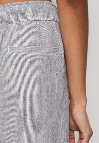 Närbild på en persons sida som bär ljusgrå byxor med tunna mörka vertikala ränder och resårmidja, som visar en bakficka och en del av deras arm.