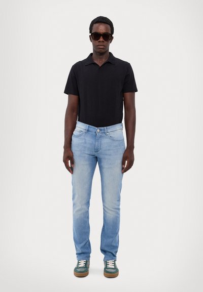 Un polo noir, un jean bleu clair et des baskets vertes avec des lacets blancs. Porté par une silhouette masculine debout, avec des lunettes de soleil.