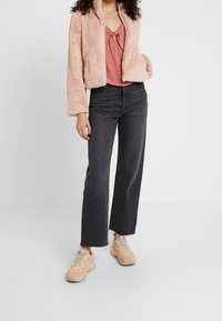 Nepbont perzikkleurig jack met ritssluiting, gecombineerd met een stofroze top, zwarte high-waisted jeans met wijde pijpen en beige sneakers.