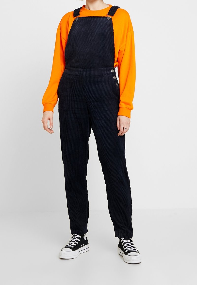 Personne portant une salopette en velours côtelé noire, un t-shirt à manches longues orange vif et des baskets montantes noires avec des semelles et lacets blancs.