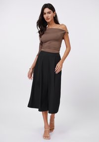 Smeđi top bez naramenica s zlatnim detaljima, u kombinaciji s crnim širokim culotte hlačama. Glatka tekstura, prilagođeni top i dizajn s naglašenim strukom.
