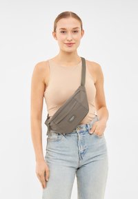 Borsa a tracolla in tessuto grigio con chiusura a zip, tracolla regolabile e tasca frontale. Indossata sopra un top beige con jeans azzurri a vita alta.